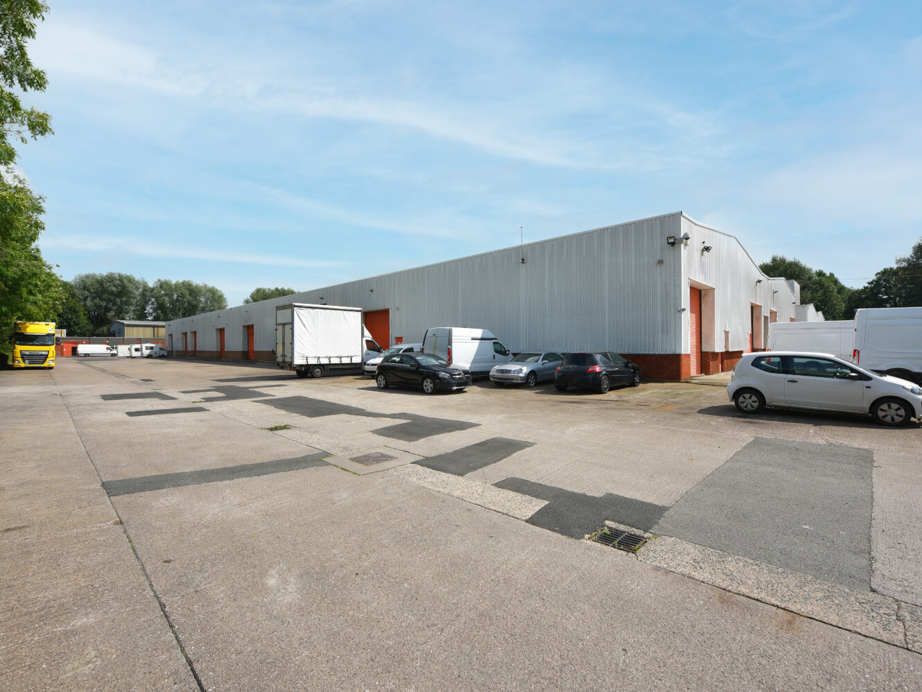 A row of industrial warehouse buildings with multiple parked vehicles and lorries in a tarmacked car park under a clear sky.