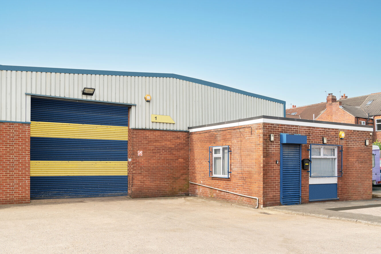 A small industrial building with a blue and yellow roller shutter door, brick walls, and a flat-roofed office section under a clear sky.