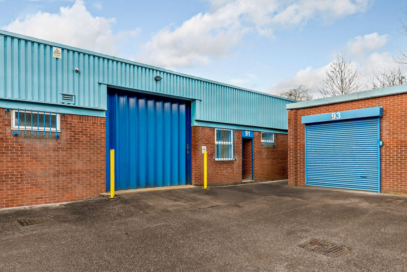 A red brick industrial building with blue metal doors, windows, and trim. Two units are labelled 91 and 93. The sky is partly cloudy.