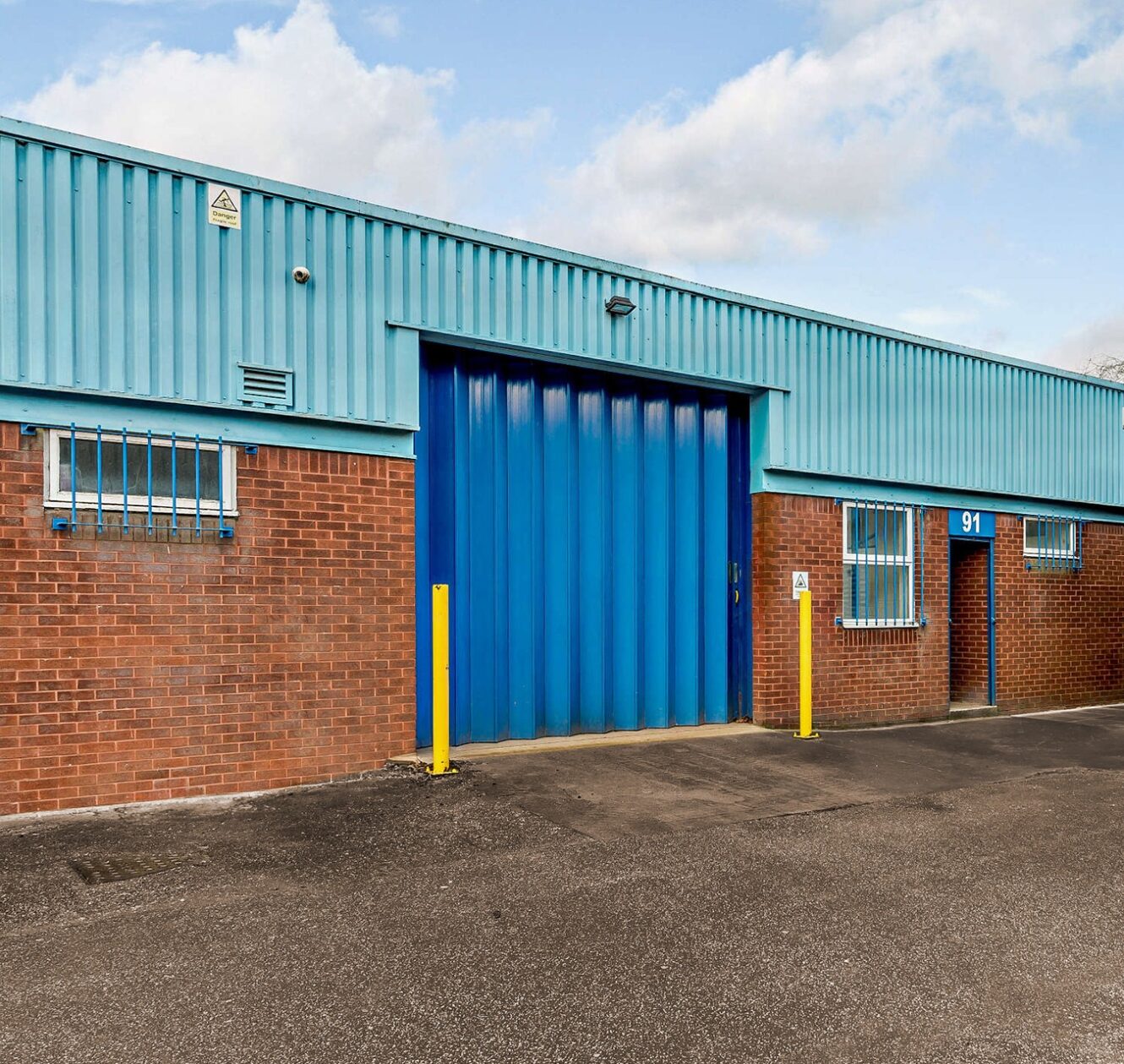 A blue industrial warehouse with a large blue roller shutter, brick lower walls, barred windows, and the number 91 displayed above a small side door.