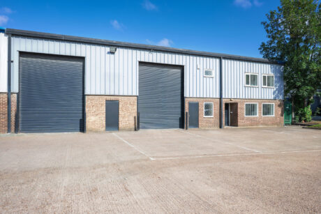 Industrial warehouse with two large roller shutters, brick lower exterior, metal upper cladding, and a small car park in front under a clear blue sky.