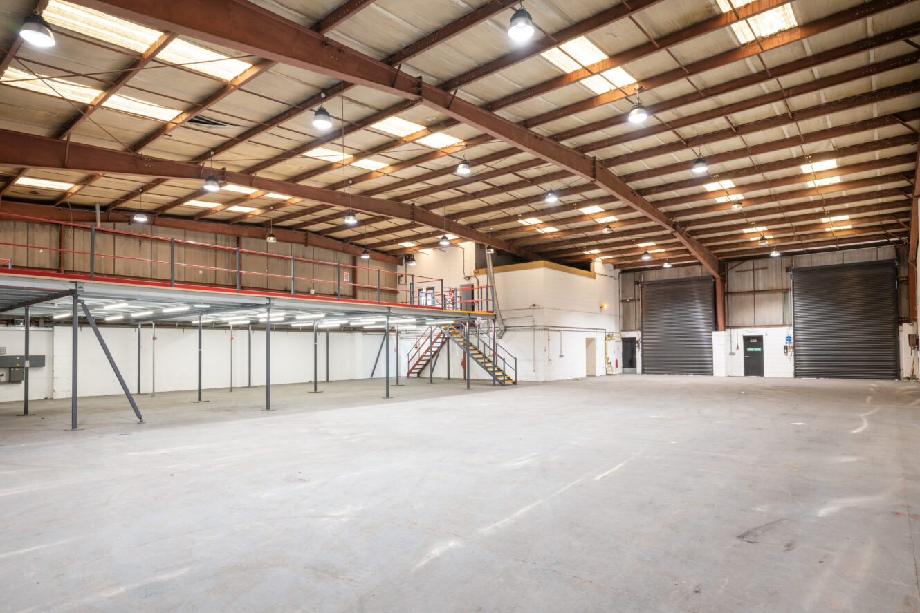 Large empty warehouse with high ceilings, exposed beams, overhead lighting, two large roller shutters, and a mezzanine level accessible by stairs.