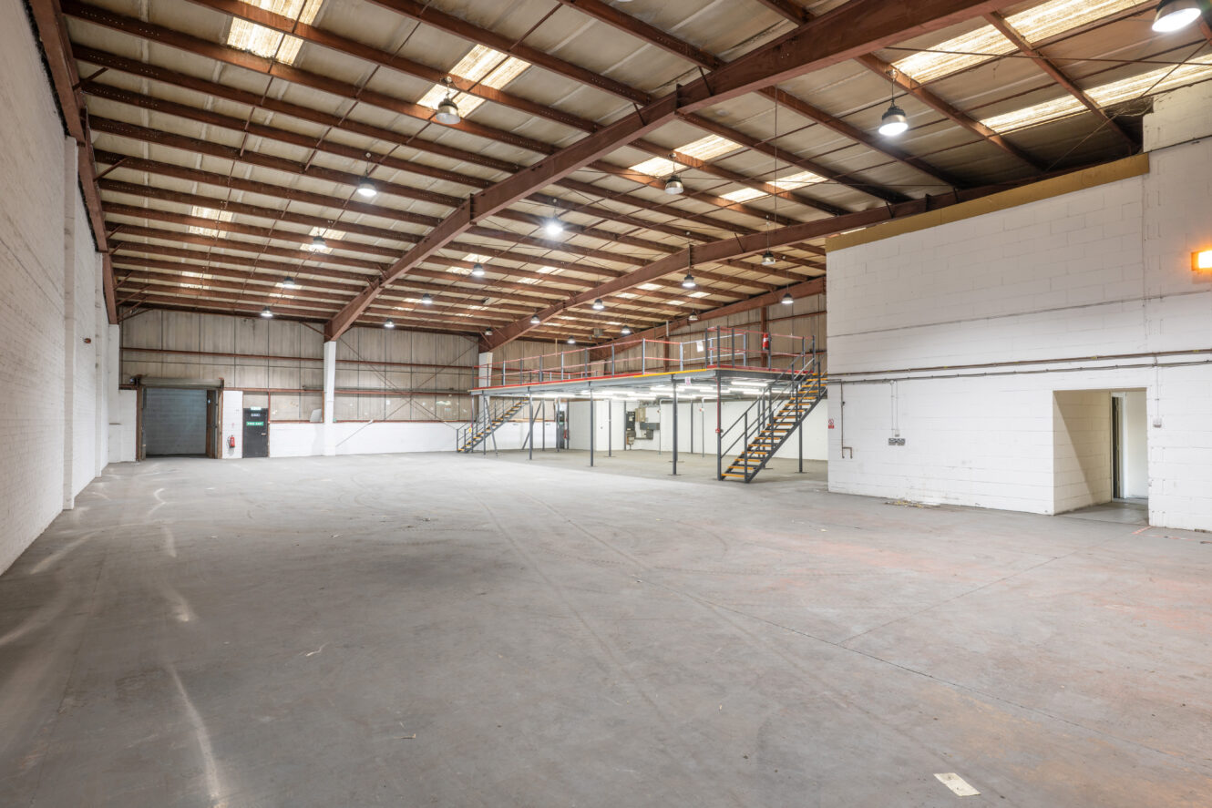 Spacious empty warehouse with high ceilings, exposed beams, concrete floor, overhead lights, staircases, and a mezzanine level.