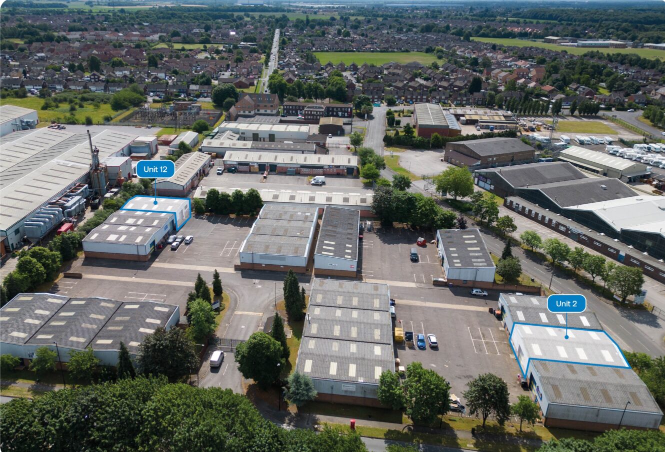 Aerial view of an industrial estate with multiple warehouse units, trees, parking spaces, and two units labelled Unit 12 and Unit 2 in blue. Residential area visible in the background.