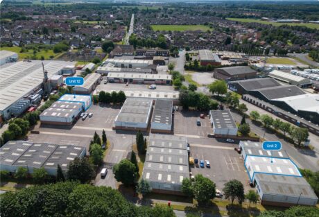 Aerial view of an industrial estate with multiple warehouse units, trees, parking spaces, and two units labelled Unit 12 and Unit 2 in blue. Residential area visible in the background.