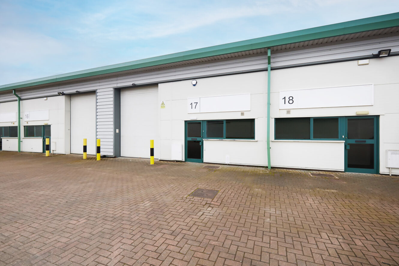 Exterior view of two industrial warehouse units numbered 17 and 18, with loading bays, white walls, and green trim, on a paved car park under a partly cloudy sky.