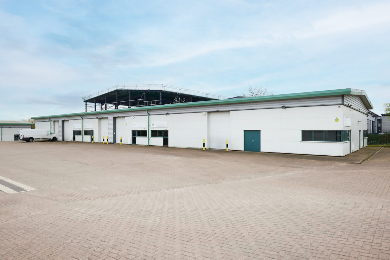 A large, white industrial warehouse building with a flat roof and several loading bay doors, set in a spacious, empty paved car park.
