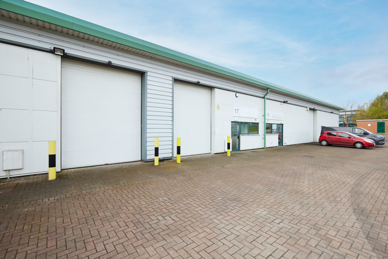 A row of industrial warehouse units with large white doors, yellow bollards, and a red car parked on a paved area in front under a clear sky.