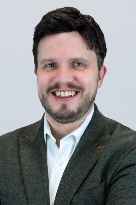 A man with dark hair and a beard is smiling, wearing a white shirt and a dark blazer, against a plain light background.