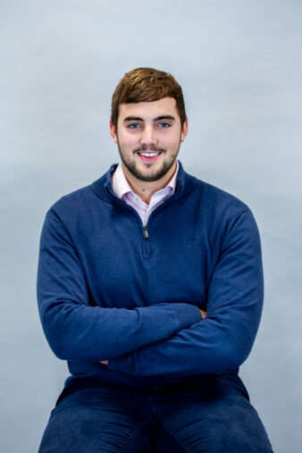 A young man with short brown hair, wearing a blue jumper and a light pink collared shirt, sits on a stool with his arms crossed against a plain light grey background.