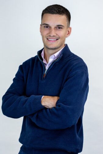 A man with short brown hair stands against a plain background, smiling, with his arms folded, wearing a navy blue jumper over a light-coloured shirt.