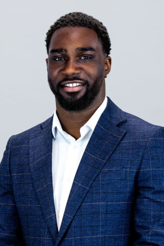 A man with short curly hair and a beard, wearing a blue checked suit jacket over a white shirt, stands against a plain light background and smiles at the camera.