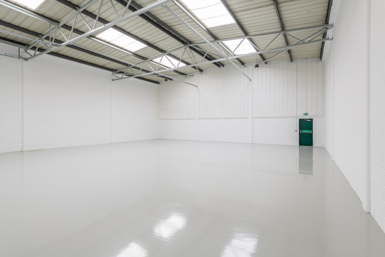 Empty industrial warehouse space with white walls, high ceiling, exposed metal beams, skylights, and a single green fire exit door.