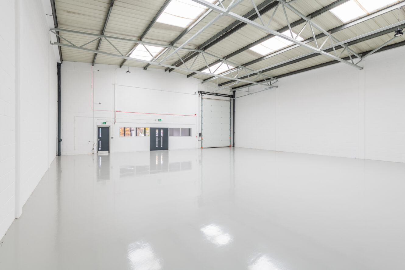 An empty industrial warehouse with white walls, a polished concrete floor, high ceiling, overhead lights, and large loading doors.