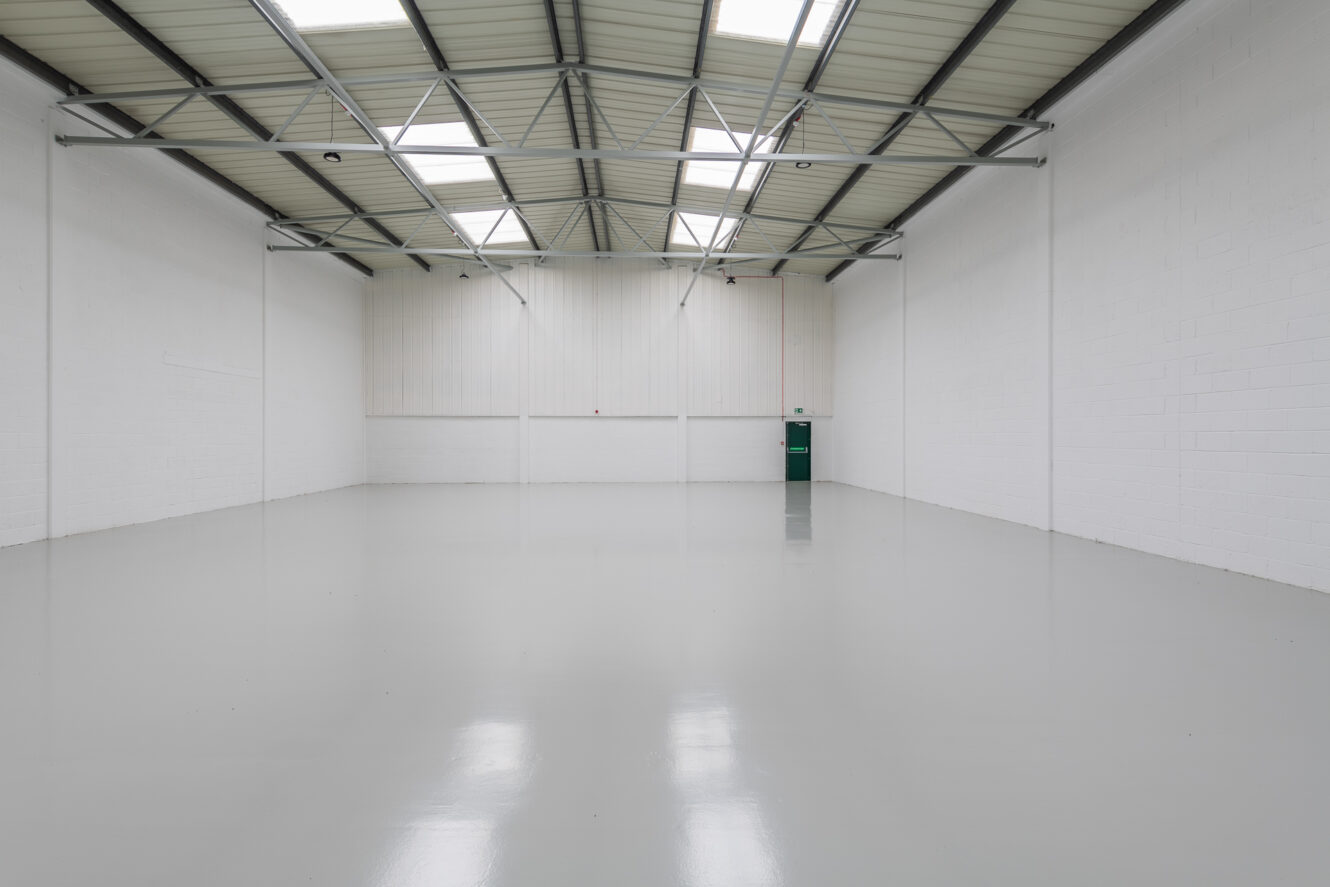 Empty industrial warehouse with white walls, clean polished floor, high ceiling, exposed beams, skylights, and a green door at the far end.