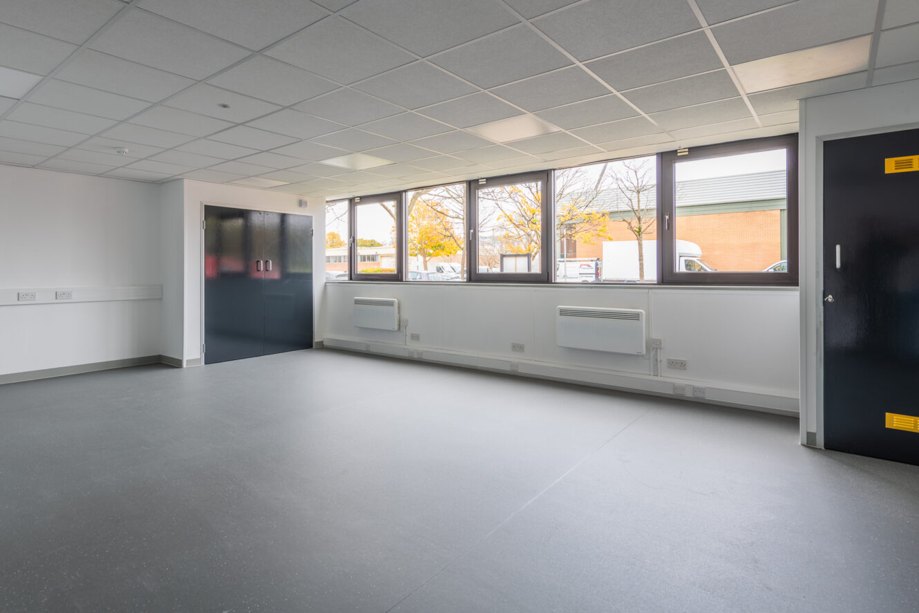 An empty, modern room with grey flooring, white walls, ceiling tiles, large windows, and dark double doors. Natural light enters through the windows, and radiators are mounted below them.