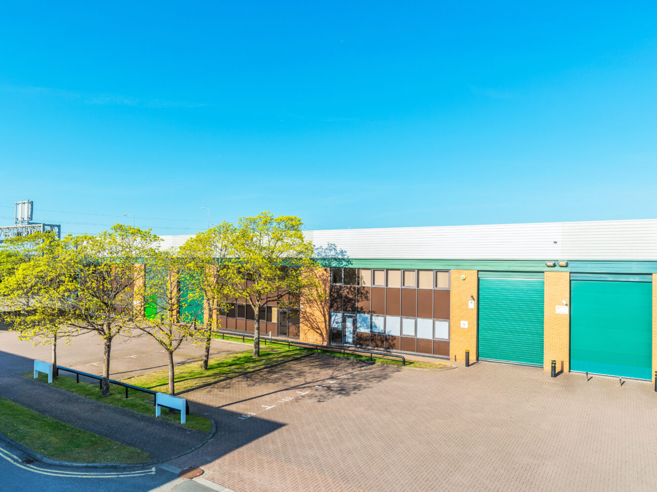 A modern industrial warehouse building with large green doors, glass windows, and a paved car park, surrounded by a few small trees under a clear blue sky.