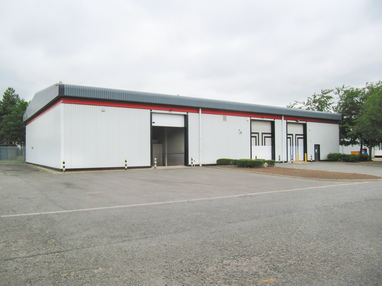 A large, white industrial warehouse with red and black trim, shown from the outside with closed and open loading bay doors and a mostly empty car park.