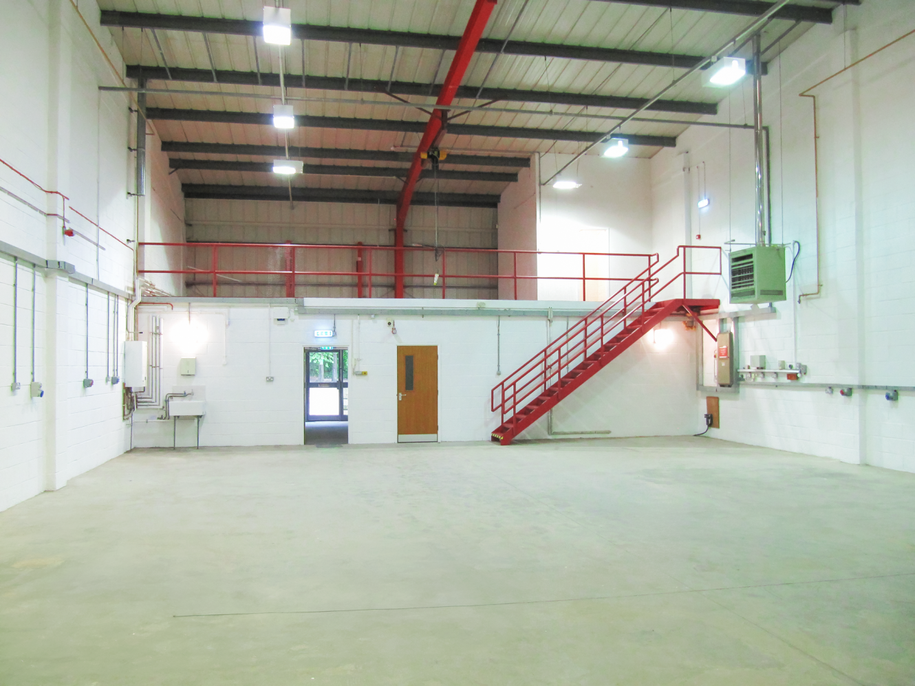 Interior of an empty industrial warehouse with white walls, a concrete floor, metal staircase to a mezzanine, and exposed pipes and lighting on the ceiling.
