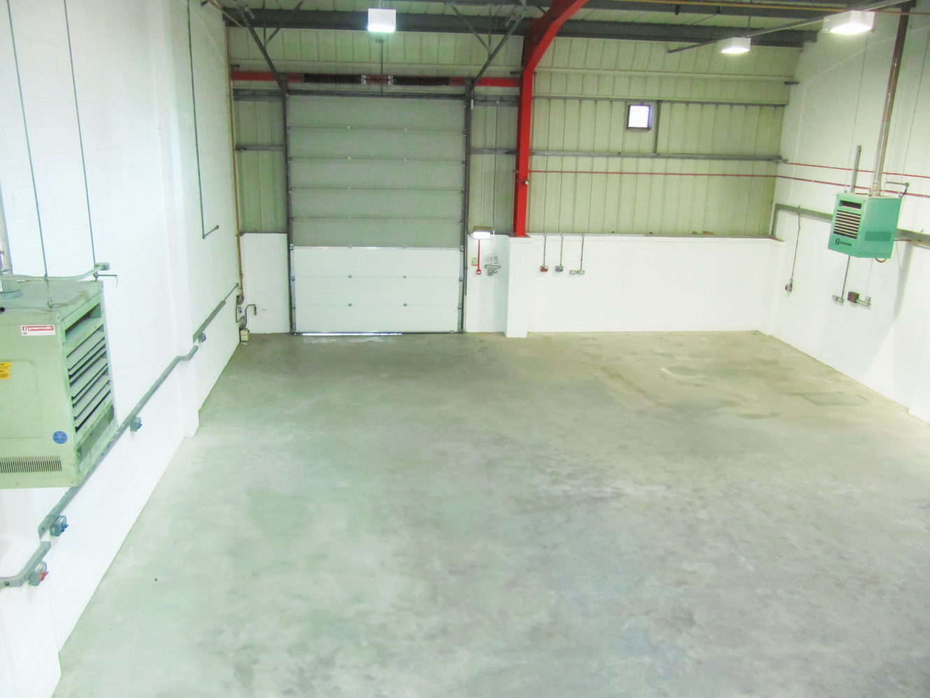 Empty industrial warehouse with concrete floor, white walls, exposed pipes, overhead lights, a large roller shutter door, and a small window on the back wall.