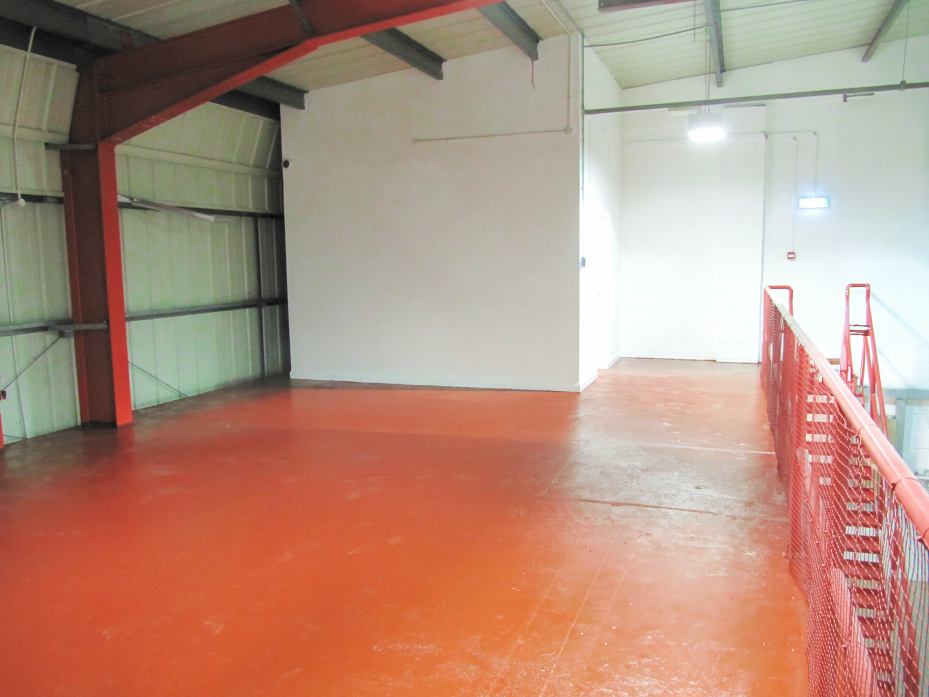 Empty industrial mezzanine with red painted floor, white walls, red railings, and exposed metal beams under a metal roof.