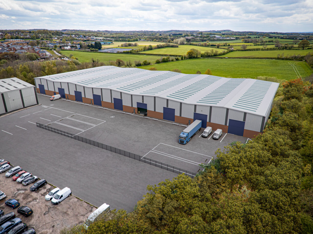 A large industrial warehouse complex with loading bays, parked lorries, and adjacent car park, surrounded by green fields and trees under a cloudy sky.