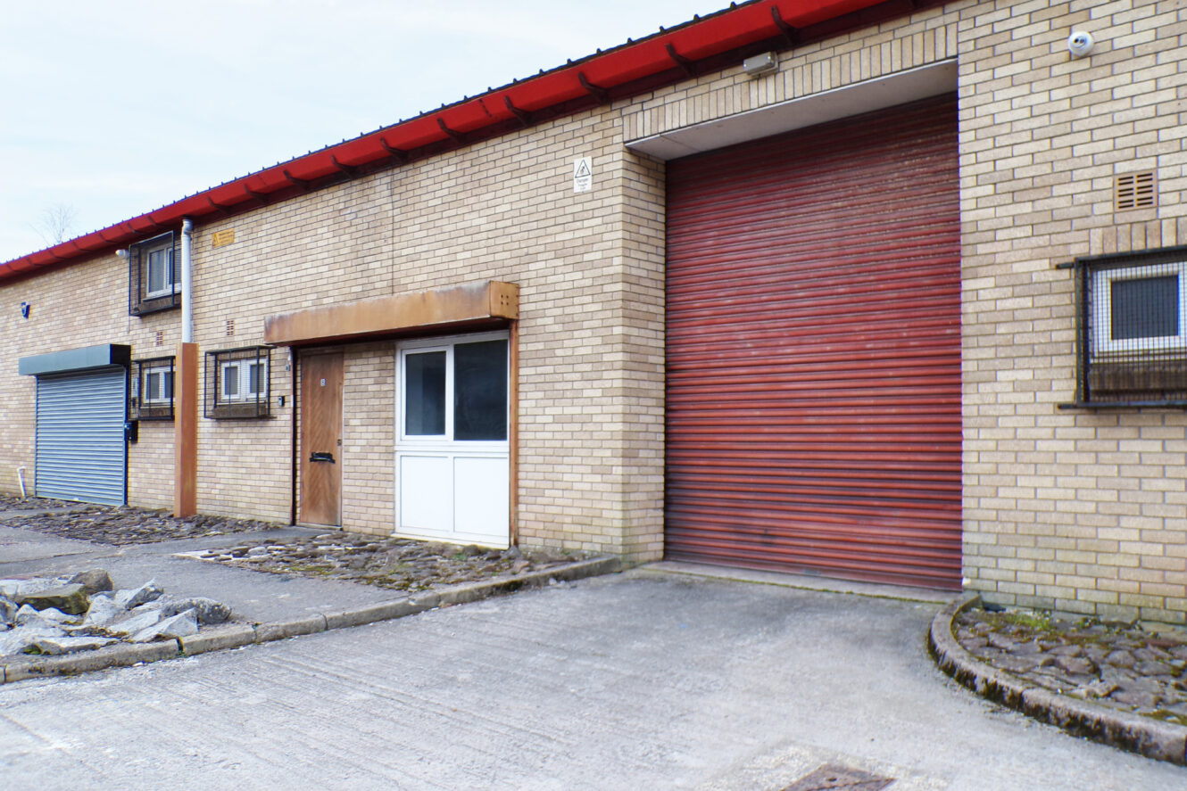 A brick industrial building with a large closed red roller shutter door, a smaller brown door, windows with security bars, and a paved driveway in front.