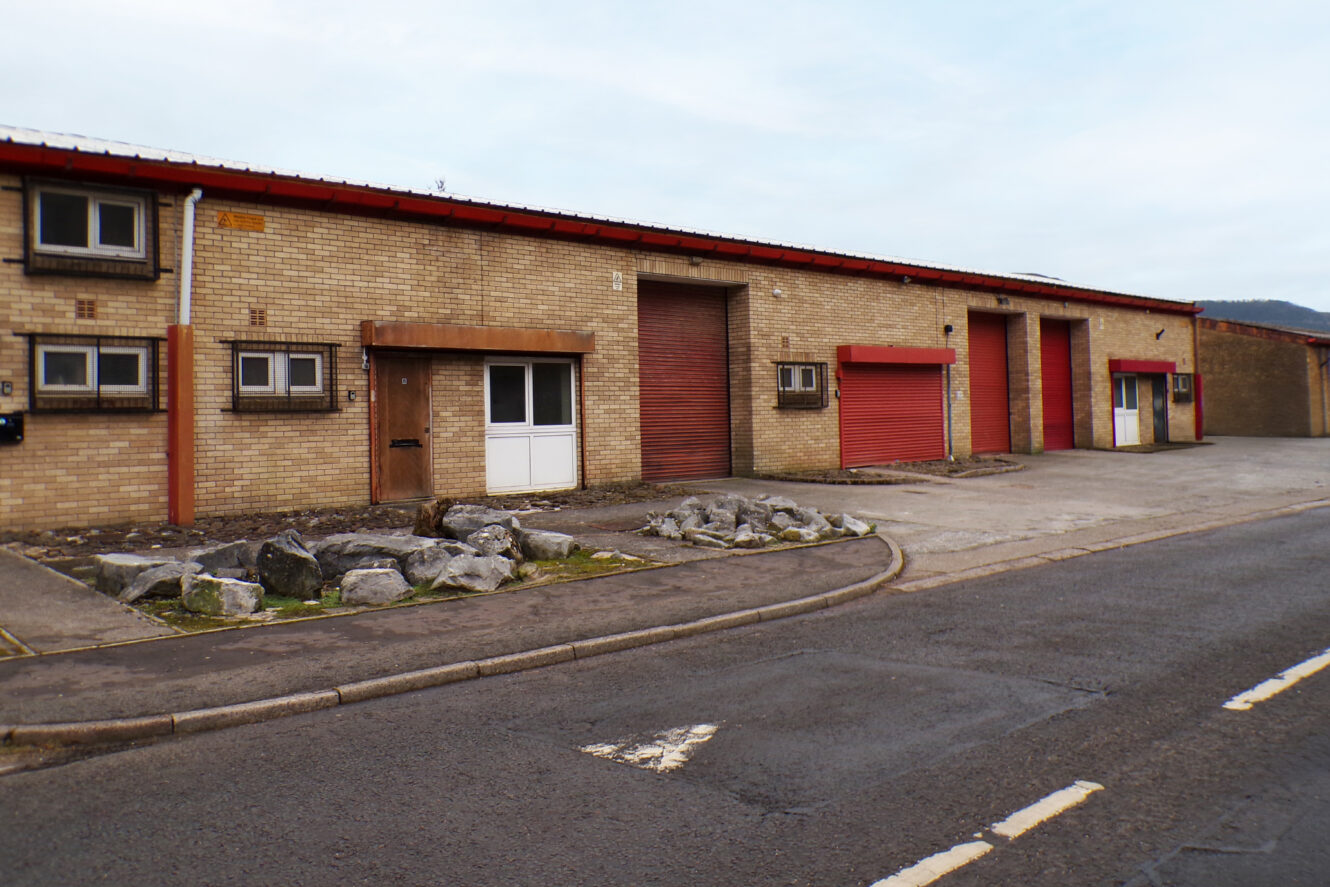 A row of industrial units with brick walls, red roller shutters, and small windows facing an empty road.