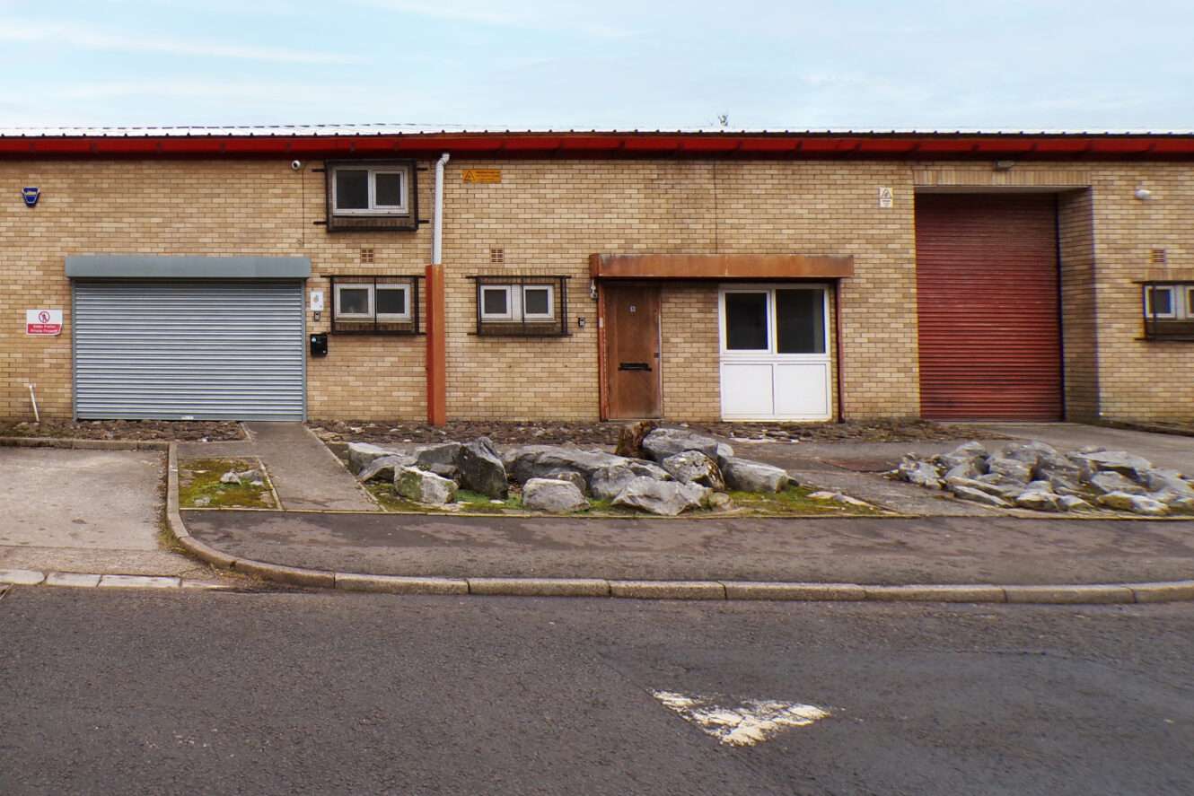 A small industrial building with beige brick walls, two metal roller shutters—one grey, one red—and a white entrance door, with rocks and gravel in the front landscaping.