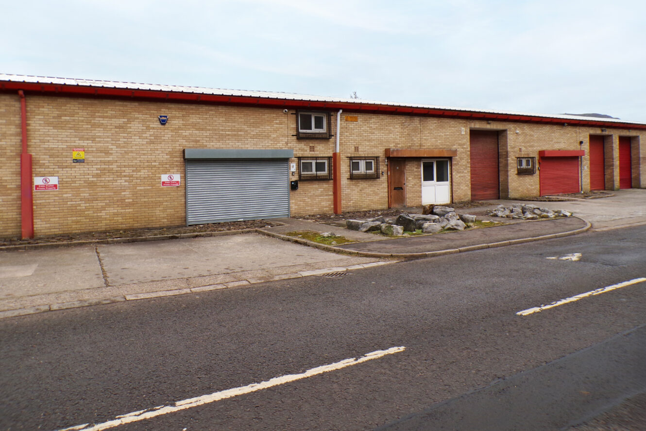 Single-storey industrial building with brick exterior, three red roller shutters, one grey shutter, multiple windows, and air conditioning units along an empty street.