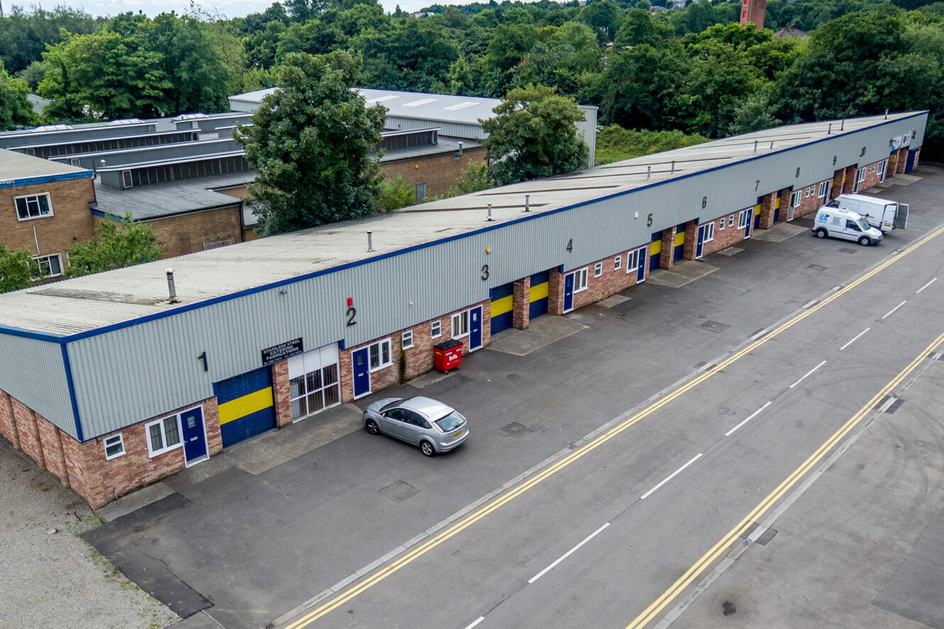 A row of industrial units with numbered doors, a grey car parked near unit 2, and a white van parked further down, surrounded by trees and neighbouring buildings.