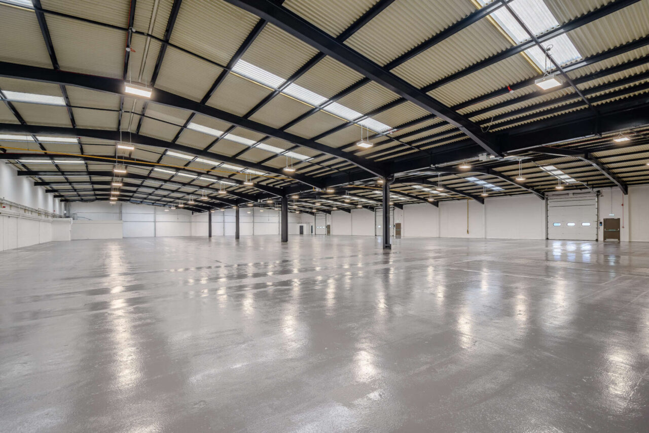 Large, empty warehouse interior with polished concrete floor, high ceilings, skylights, and black steel beams. Roller shutter doors visible at the far end.