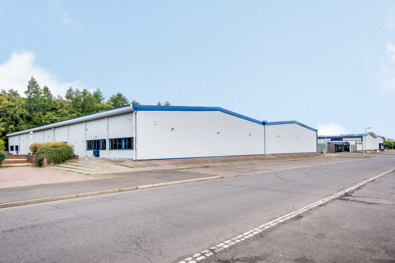 A large, light grey industrial warehouse with blue trim sits beside an empty road on a clear day.