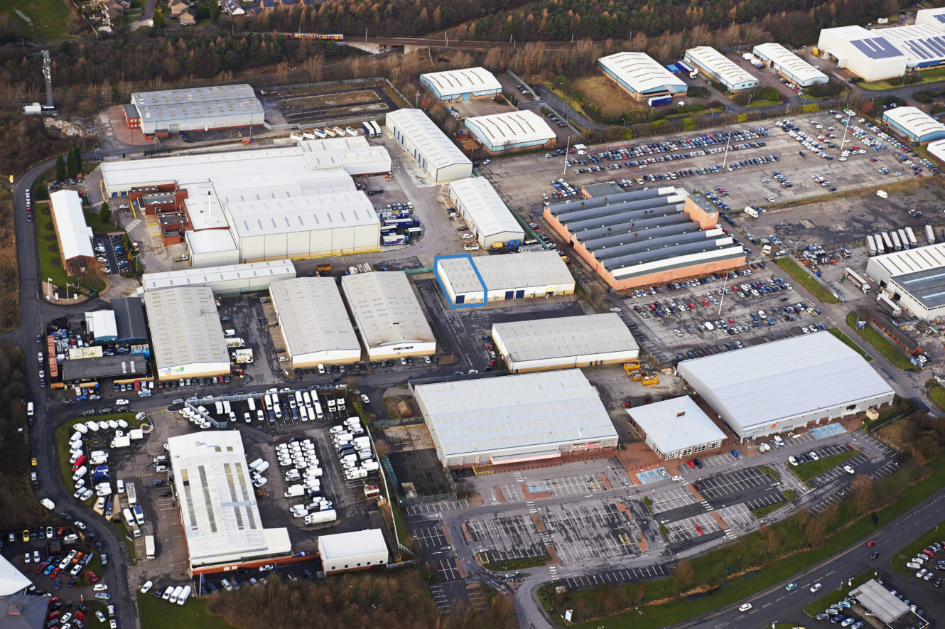 Aerial view of an industrial estate with multiple large warehouse buildings, car parks, and surrounding roads, bordered by trees.