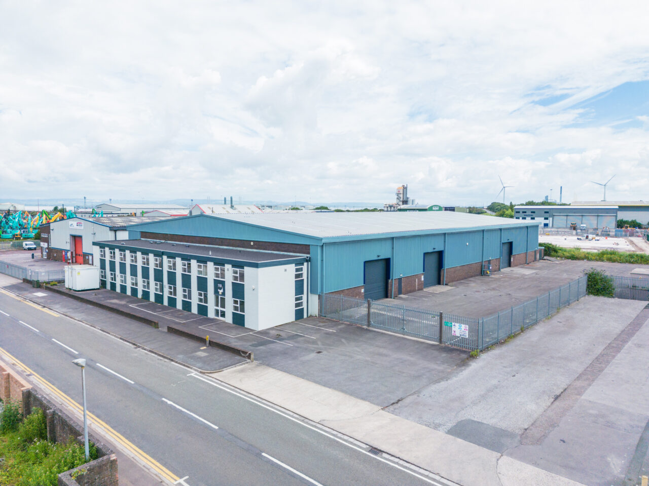 A large industrial warehouse with blue panels, multiple loading bays, and an adjacent office building, surrounded by a fenced yard, located on an empty street.