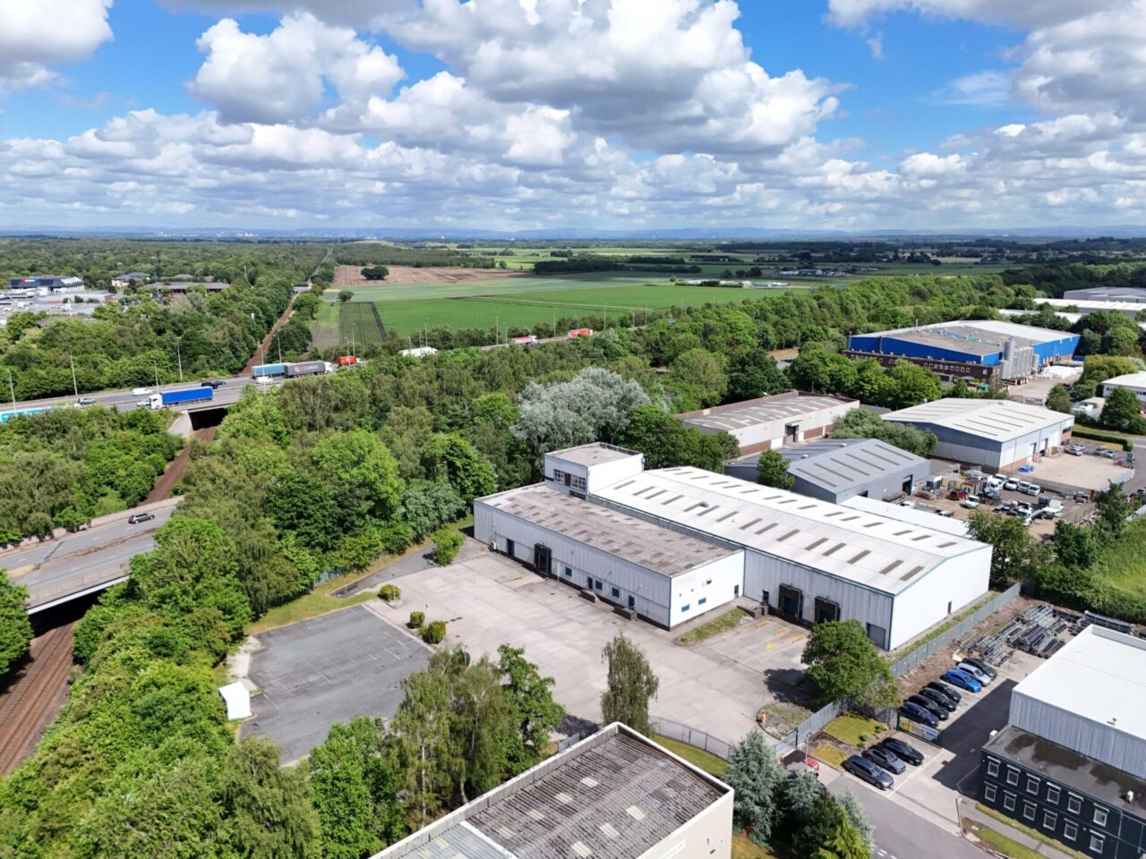 Aerial view of an industrial area with warehouses, car parks, surrounding trees, and open fields in the background under a partly cloudy sky.