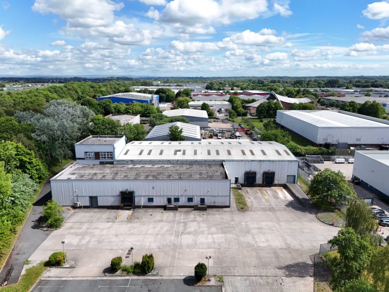 Aerial view of a large industrial warehouse complex surrounded by paved car parks, trees, and other commercial buildings under a partly cloudy sky.