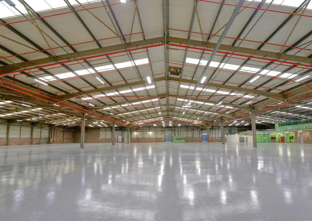 Large empty warehouse with polished concrete floors, high ceilings, exposed beams, and fluorescent lighting. Green doors and windows line the far right wall.