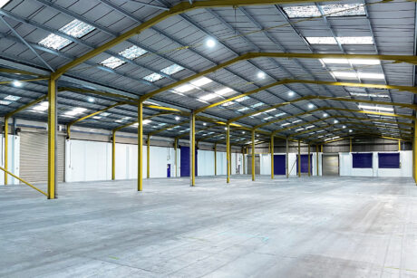 Interior of a large, empty warehouse with yellow steel beams, high ceiling, concrete floor, and skylights letting in natural light.