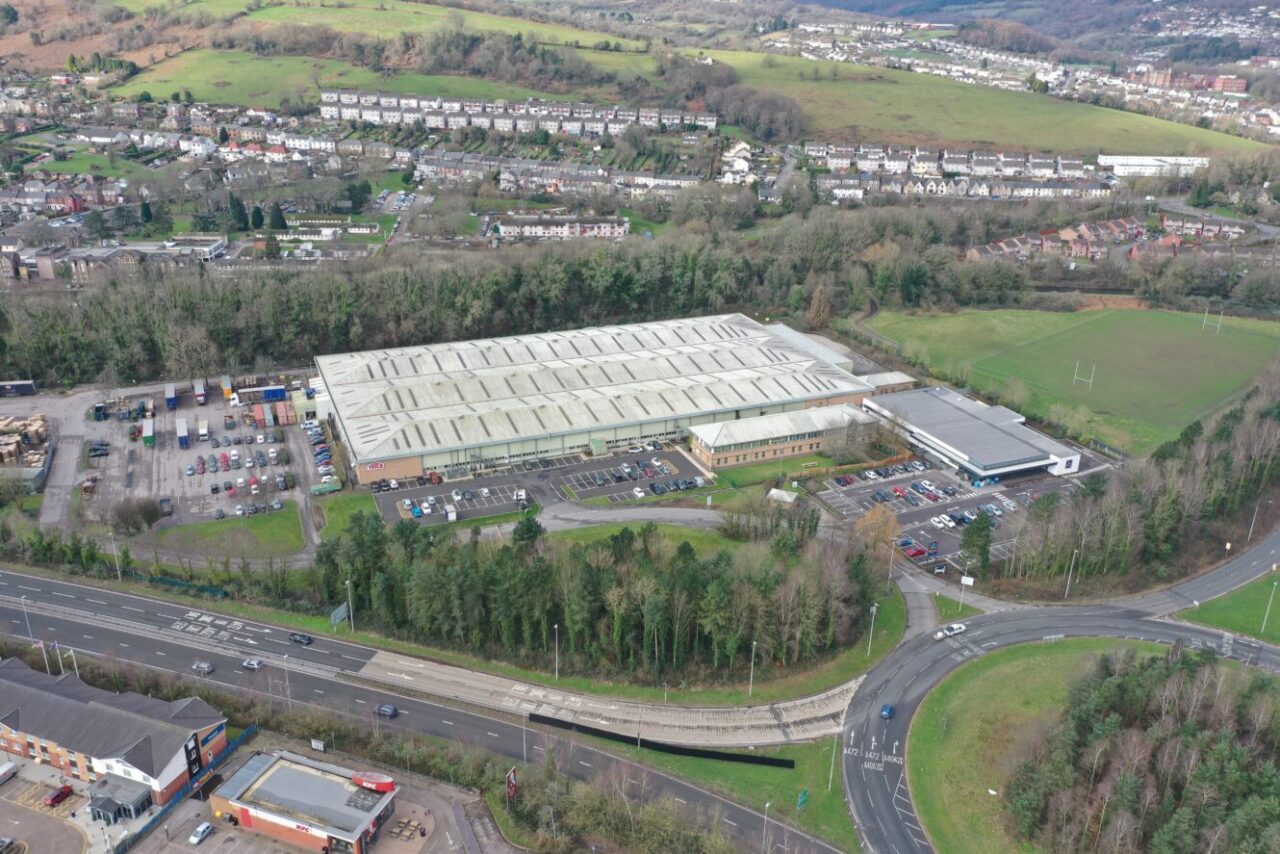 Aerial view of a large industrial building with adjacent car parks, surrounded by trees and roads, with a residential area and green hills in the background.