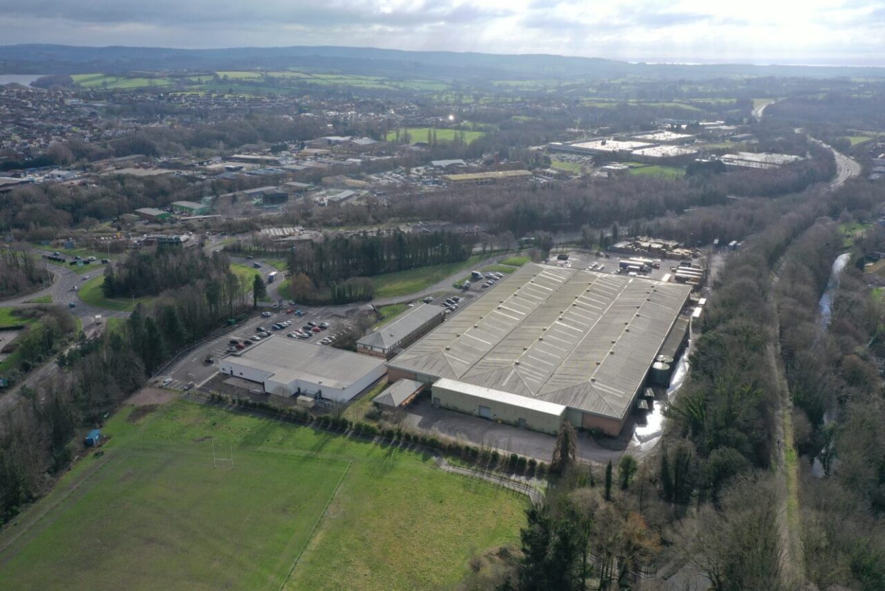 Aerial view of a large warehouse complex surrounded by trees and fields, with roads, car parks, and buildings visible in the background.