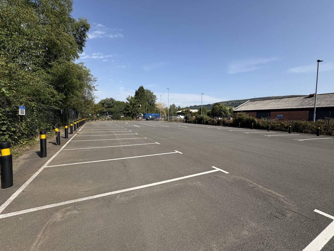 An empty outdoor car park with marked parking spaces, black and yellow bollards, trees on the left, and a building on the right under a clear sky.