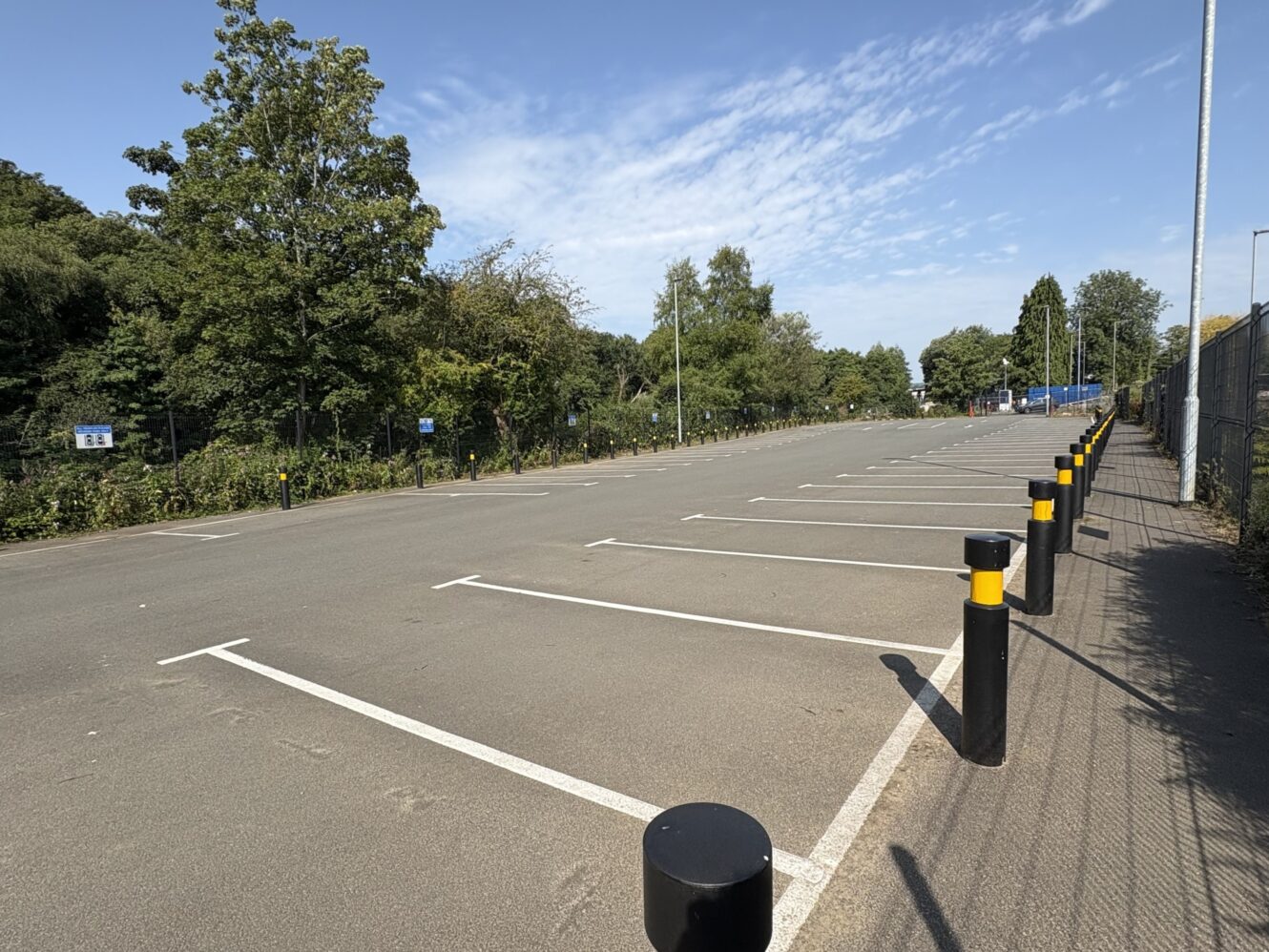 An empty outdoor car park with marked spaces, bordered by trees on one side and a fence on the other, under a clear blue sky.