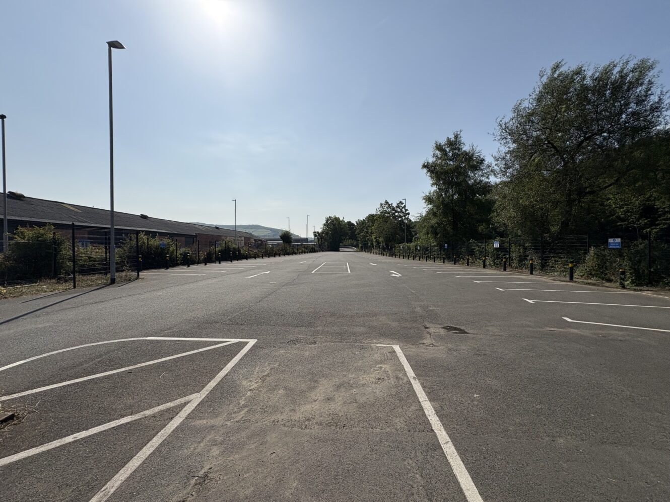 An empty outdoor car park on a clear, sunny day, with marked parking spaces and some trees and buildings in the background.