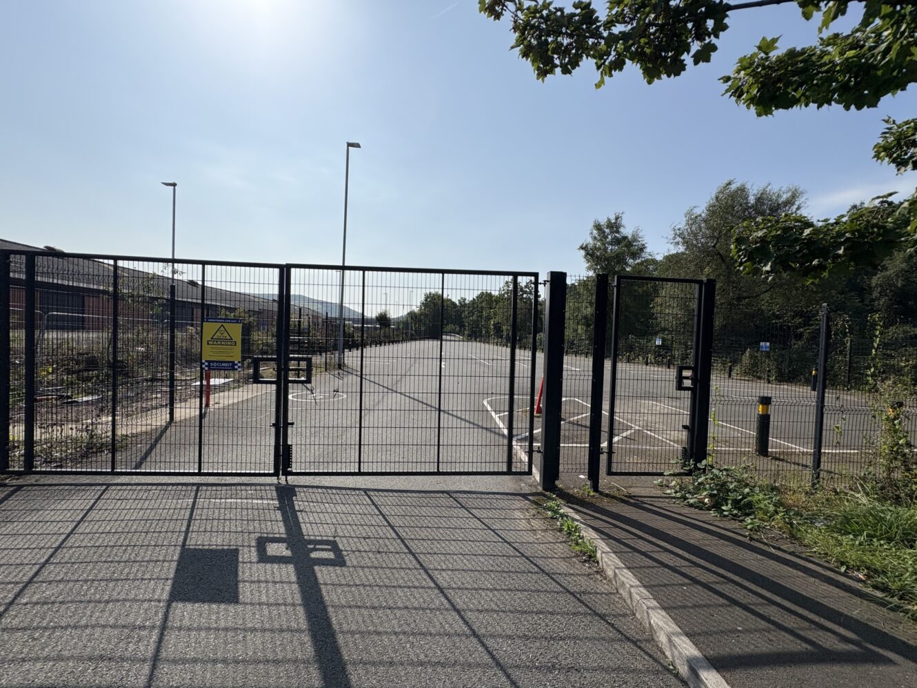 A closed metal gate blocks entry to an empty tarmac road or car park, with trees and a clear sky in the background.