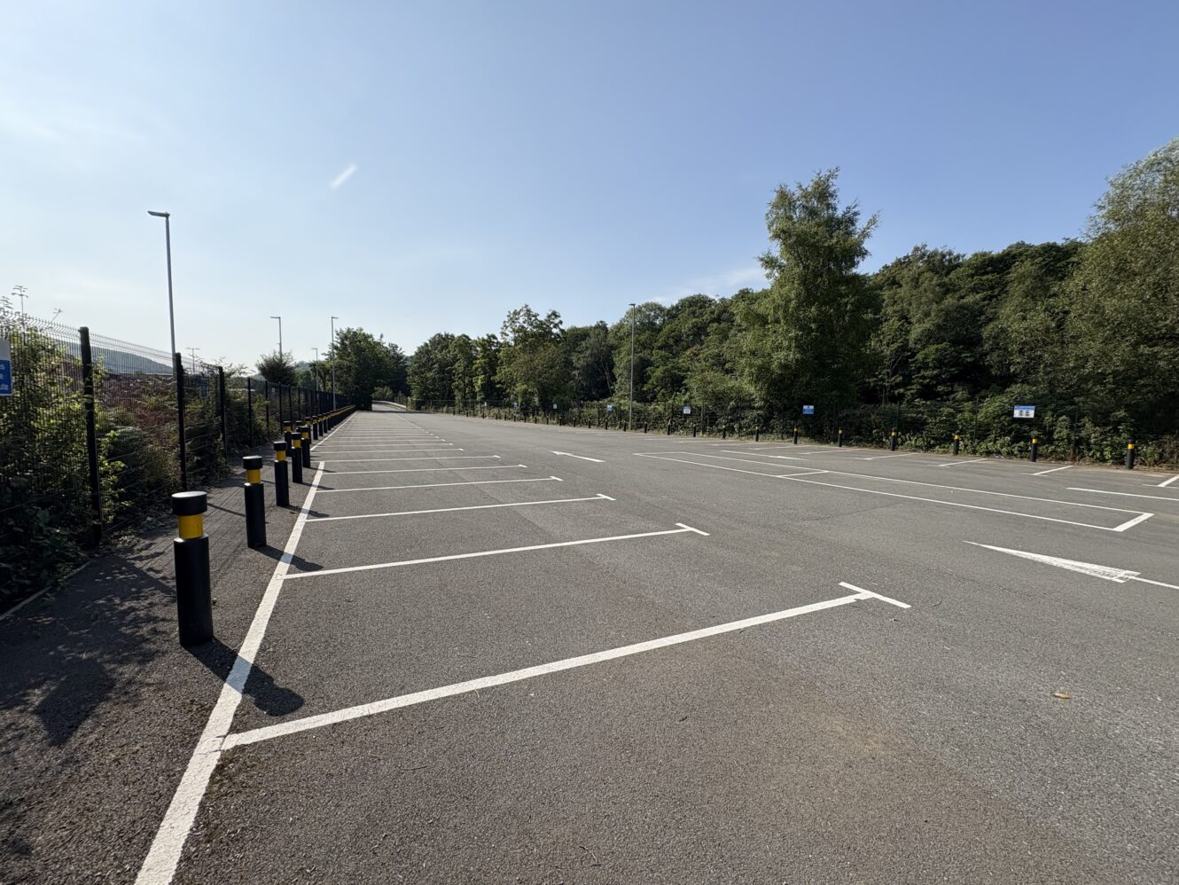 An empty outdoor car park with clearly marked parking spaces, surrounded by trees and greenery under a clear blue sky.