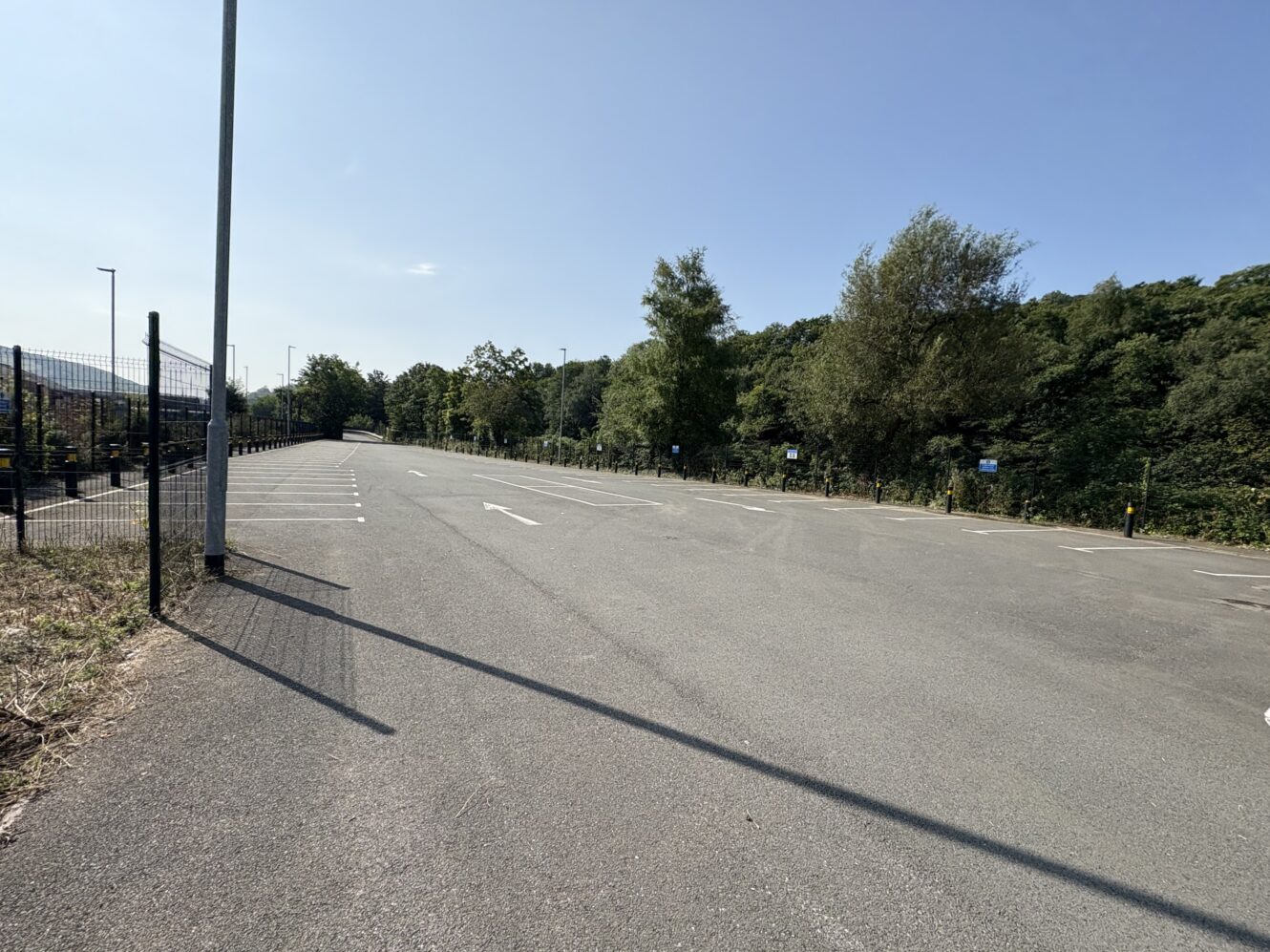 An empty outdoor car park on a sunny day, bordered by a fence on the left and trees on the right.