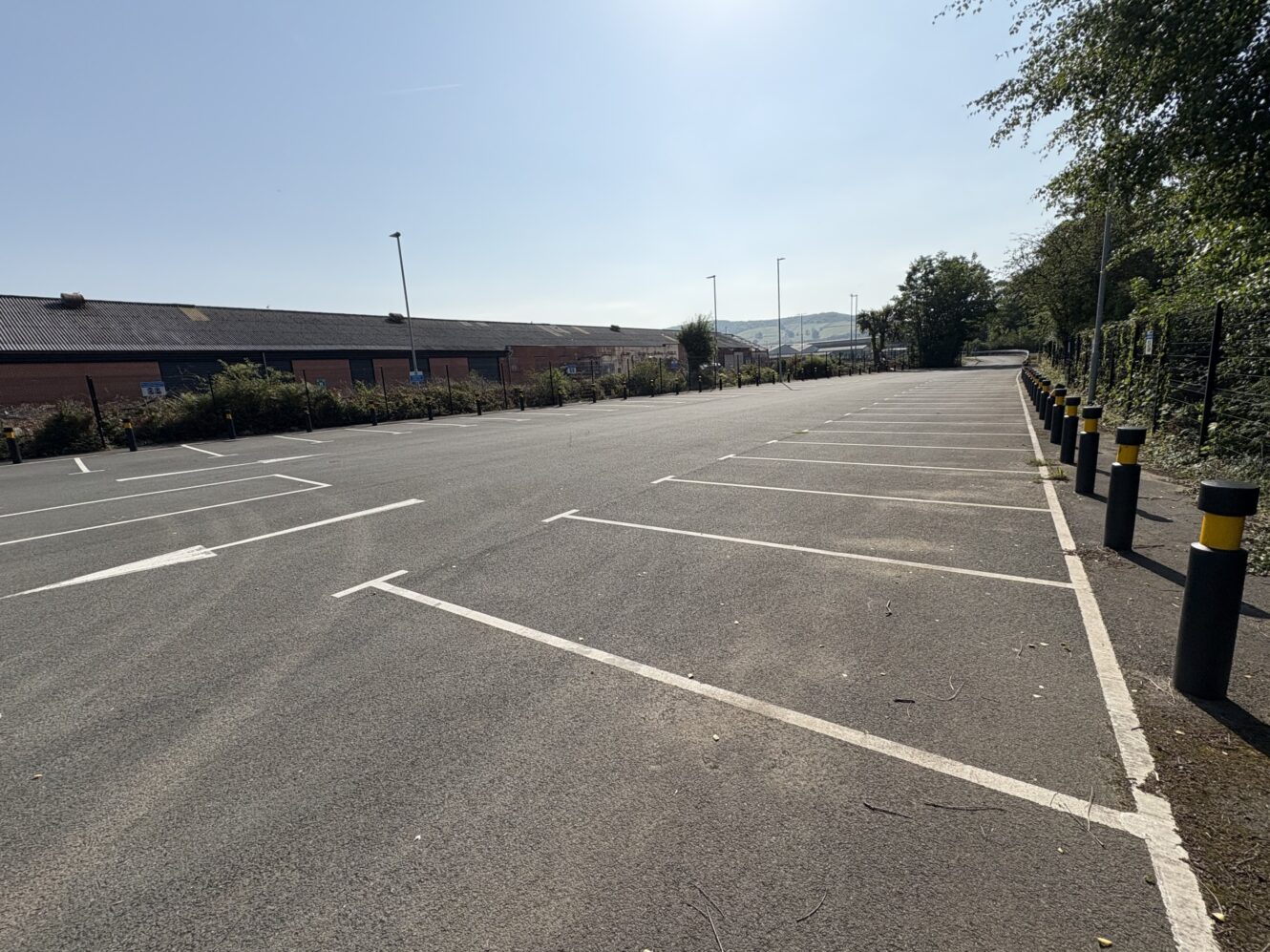 An empty outdoor car park with marked parking spaces, bordered by bollards and greenery, under a clear sky.