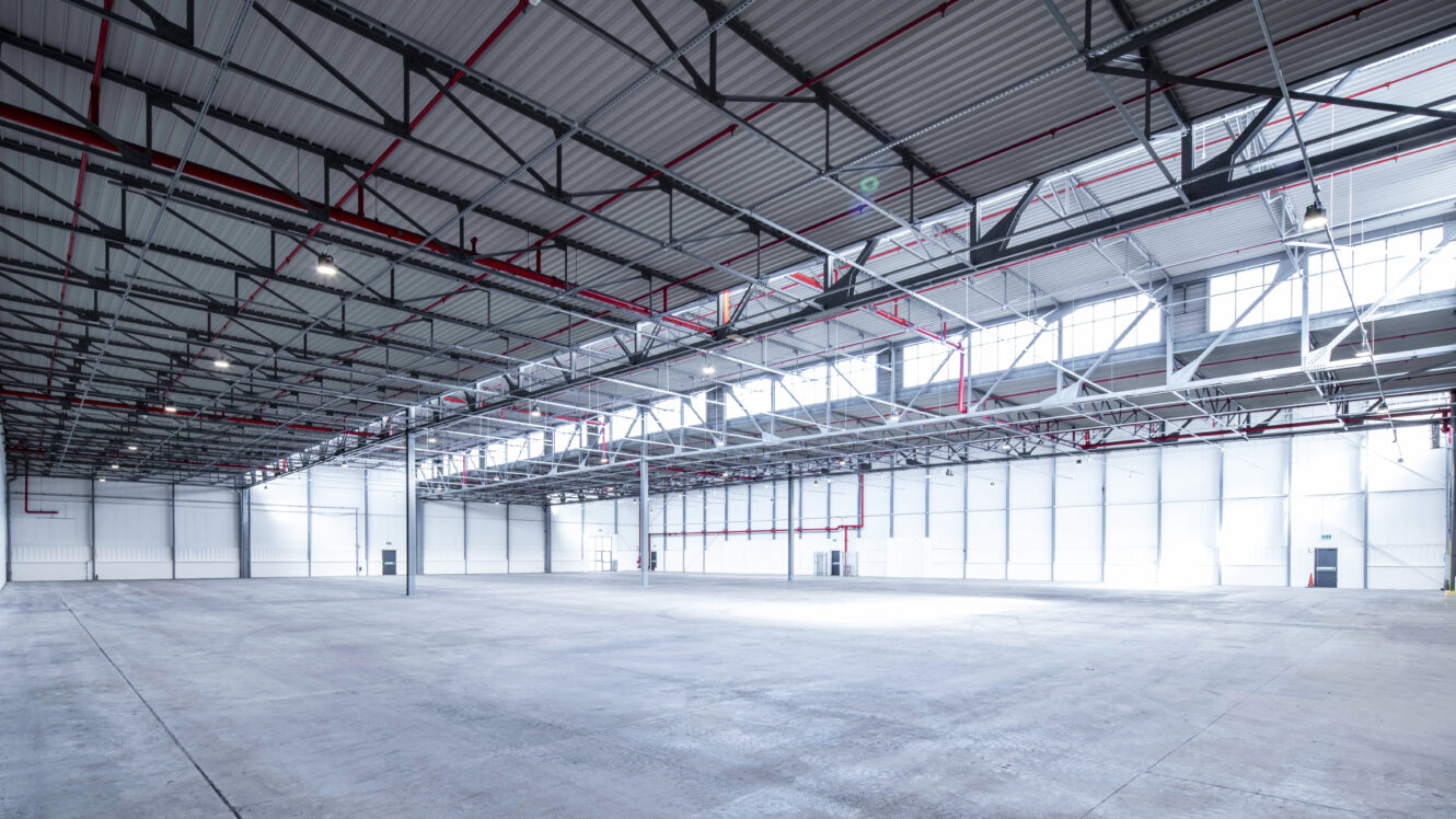 Large empty warehouse with high ceilings, exposed beams, red pipes, and windows along the upper walls allowing natural light to enter.
