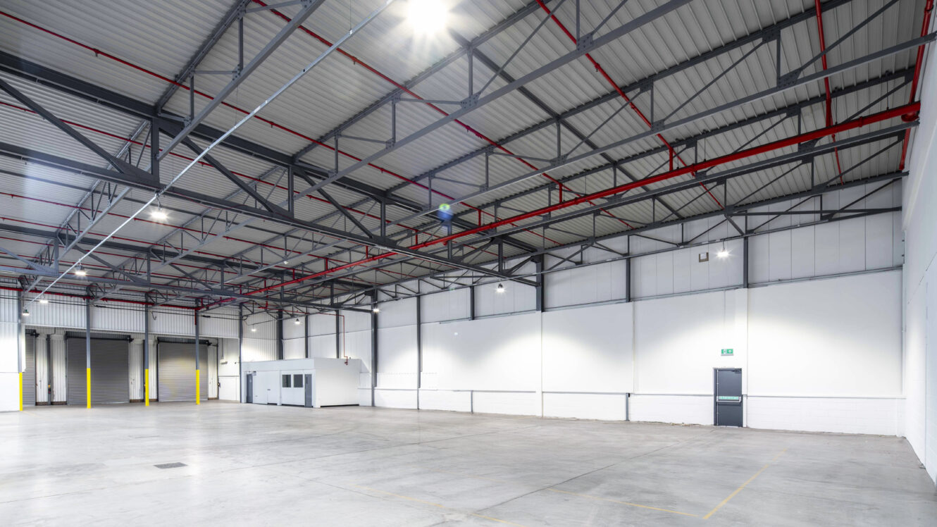 Large empty warehouse with high ceilings, visible steel beams, overhead lighting, concrete floor, loading bay doors, and a small interior office structure near the back wall.